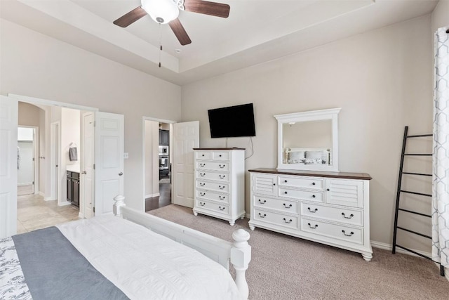 bedroom featuring light carpet, baseboards, ceiling fan, ensuite bathroom, and a tray ceiling