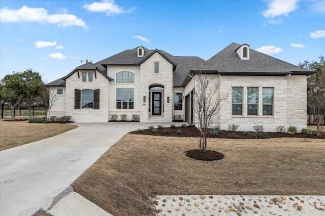 french provincial home with a shingled roof, stone siding, and fence