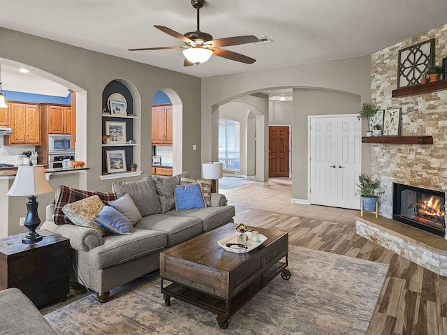 living area with visible vents, a ceiling fan, a stone fireplace, light wood-type flooring, and built in shelves