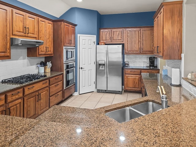 kitchen featuring brown cabinets, appliances with stainless steel finishes, a sink, dark stone countertops, and under cabinet range hood