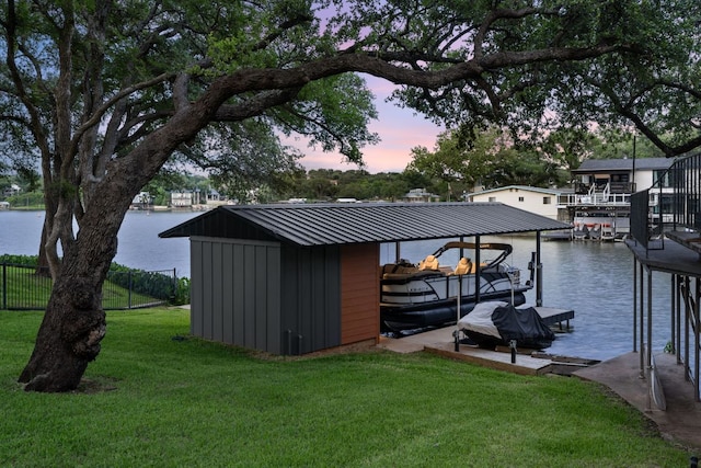 outdoor structure at dusk with a water view, a yard, boat lift, and fence