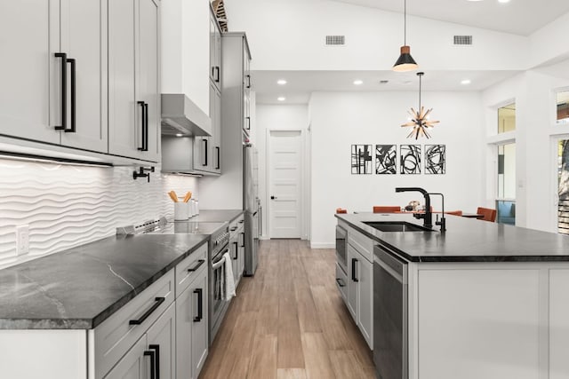 kitchen featuring a sink, wall chimney range hood, appliances with stainless steel finishes, light wood finished floors, and a center island with sink