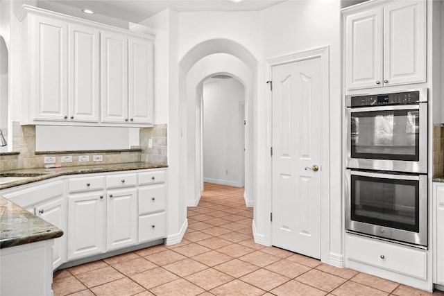 kitchen with backsplash, double oven, light tile patterned flooring, arched walkways, and white cabinetry