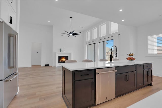 kitchen with a warm lit fireplace, stainless steel appliances, light countertops, light wood finished floors, and an island with sink