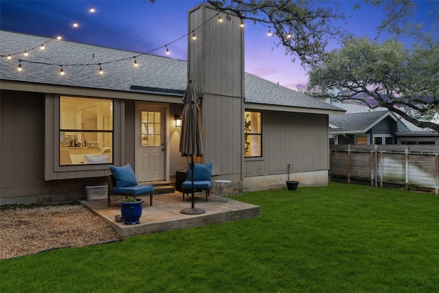 back of house at dusk with a patio, a yard, a chimney, a shingled roof, and fence