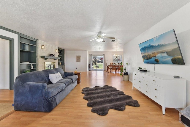 living area with a textured ceiling, light wood-type flooring, a brick fireplace, built in shelves, and ceiling fan with notable chandelier