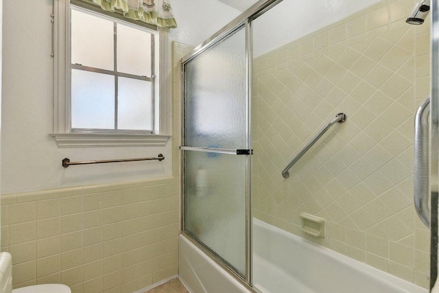 bathroom featuring toilet, shower / bath combination with glass door, tile walls, and a wainscoted wall