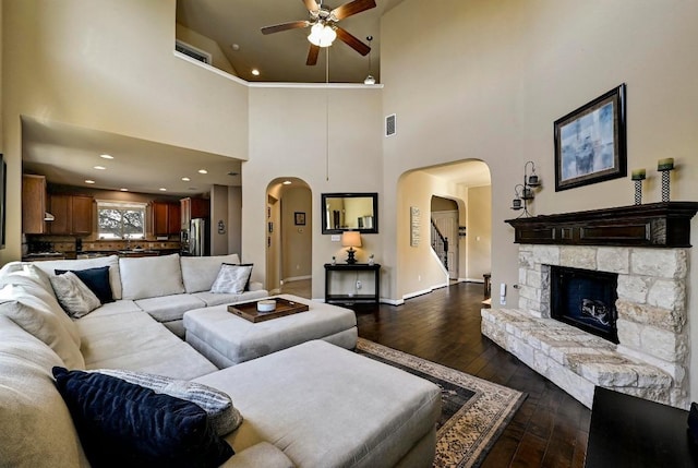 living area featuring arched walkways, a stone fireplace, dark wood-style flooring, a ceiling fan, and visible vents