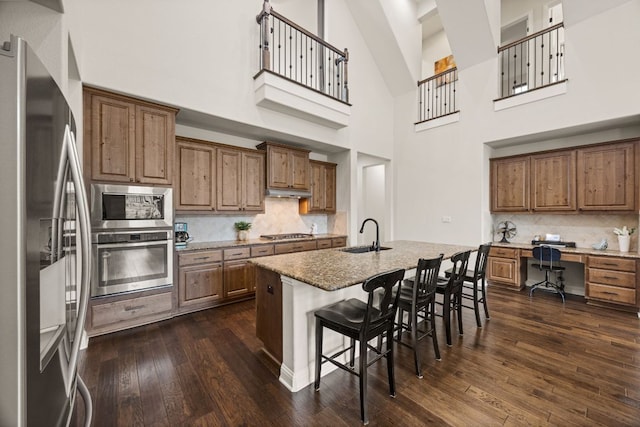 kitchen with appliances with stainless steel finishes, brown cabinets, dark wood-style flooring, light stone countertops, and a sink