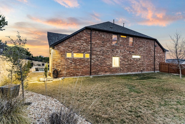 property exterior at dusk with a yard, brick siding, roof with shingles, and fence