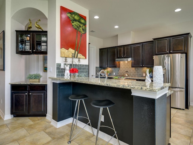 kitchen with tasteful backsplash, a breakfast bar area, freestanding refrigerator, under cabinet range hood, and a sink