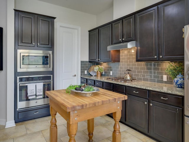 kitchen with light tile patterned floors, under cabinet range hood, appliances with stainless steel finishes, light stone countertops, and tasteful backsplash