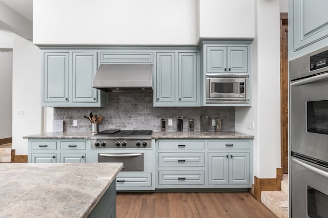 kitchen featuring tasteful backsplash, appliances with stainless steel finishes, wood finished floors, light stone countertops, and under cabinet range hood