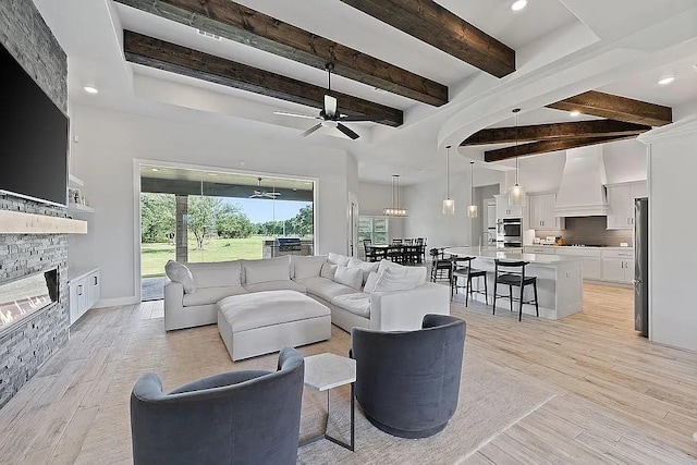 living area featuring a stone fireplace, light wood-type flooring, beam ceiling, and a ceiling fan