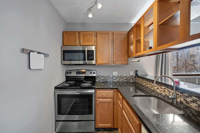 kitchen featuring stainless steel appliances, brown cabinetry, glass insert cabinets, a sink, and dark stone counters