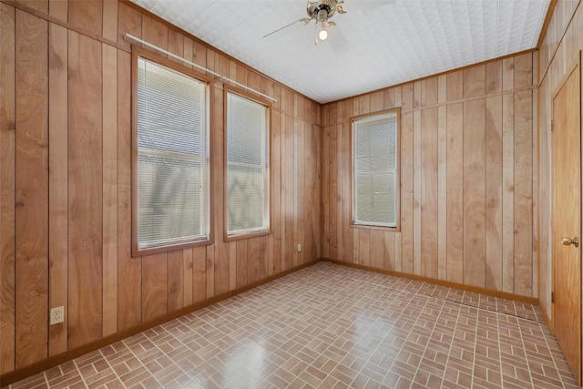 unfurnished room featuring brick floor, wooden walls, baseboards, and a ceiling fan