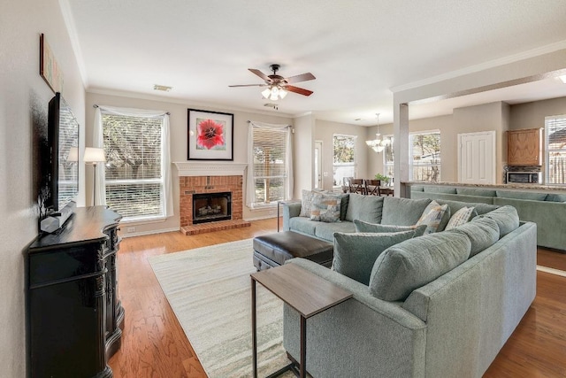 living room featuring visible vents, ornamental molding, wood finished floors, a brick fireplace, and ceiling fan with notable chandelier