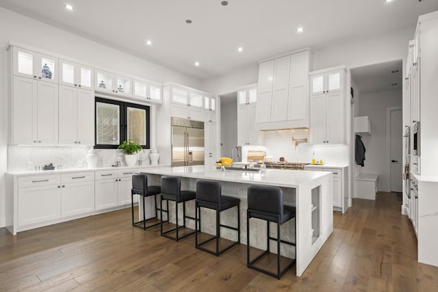 kitchen with built in fridge, a sink, white cabinets, an island with sink, and dark wood finished floors