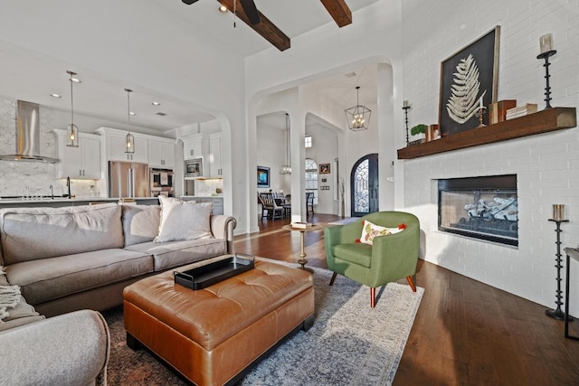 living room with arched walkways, dark wood finished floors, beamed ceiling, a high ceiling, and a brick fireplace