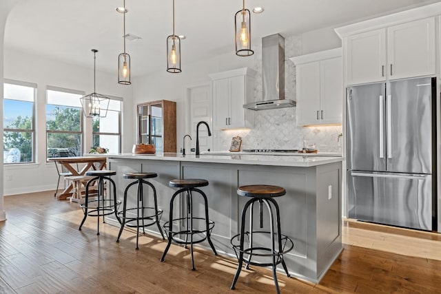 kitchen with backsplash, freestanding refrigerator, an island with sink, wood finished floors, and wall chimney exhaust hood