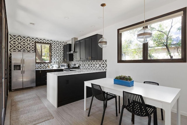 kitchen with light countertops, backsplash, wall chimney range hood, stainless steel fridge, and a peninsula