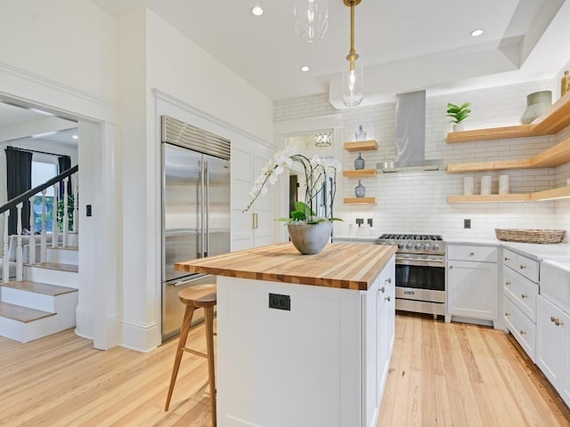 kitchen featuring wall chimney exhaust hood, high quality appliances, open shelves, and butcher block countertops