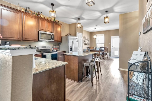 kitchen with light wood finished floors, brown cabinetry, appliances with stainless steel finishes, a center island, and light stone countertops