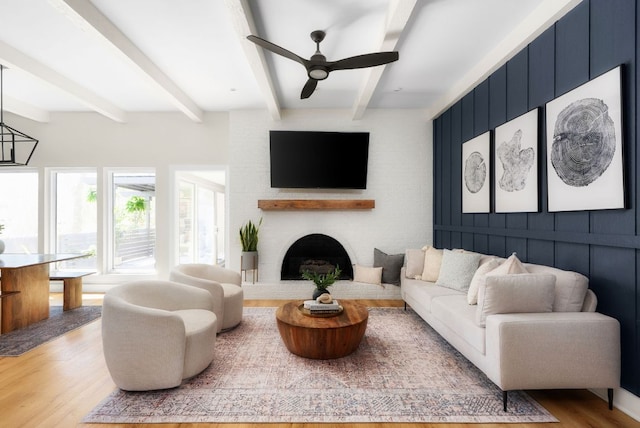 living area with ceiling fan, a brick fireplace, beamed ceiling, and wood finished floors