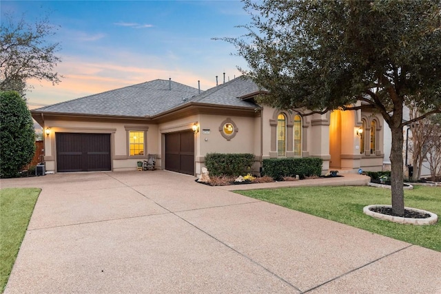 view of front facade with a garage, driveway, a lawn, roof with shingles, and stucco siding