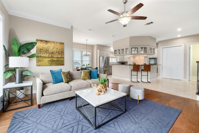 living room with ornamental molding, visible vents, light wood-style flooring, and ceiling fan with notable chandelier