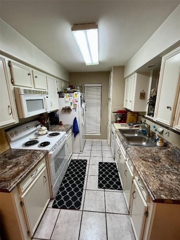 kitchen featuring dark countertops, light tile patterned flooring, a sink, white cabinets, and white appliances