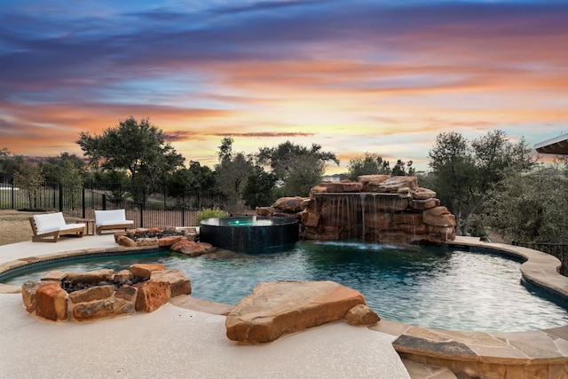 pool at dusk featuring a pool with connected hot tub and fence