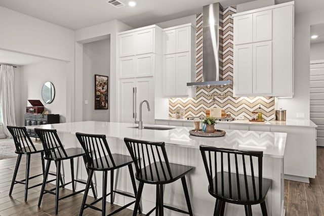 kitchen featuring dark wood finished floors, an island with sink, light countertops, wall chimney range hood, and a sink