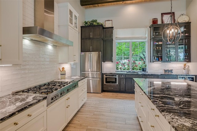 kitchen featuring a chandelier, wall chimney range hood, appliances with stainless steel finishes, dark stone counters, and light wood finished floors