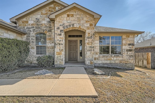 doorway to property with a shingled roof, stone siding, and fence