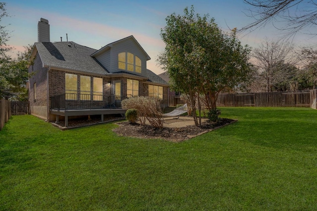 back of house at dusk with a yard, a fenced backyard, a chimney, a deck, and brick siding