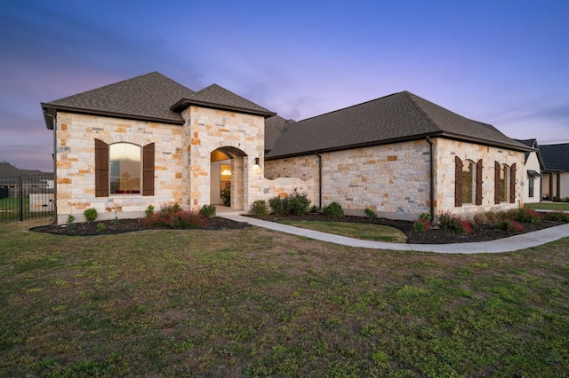 french country home with a front lawn, fence, stone siding, and a shingled roof