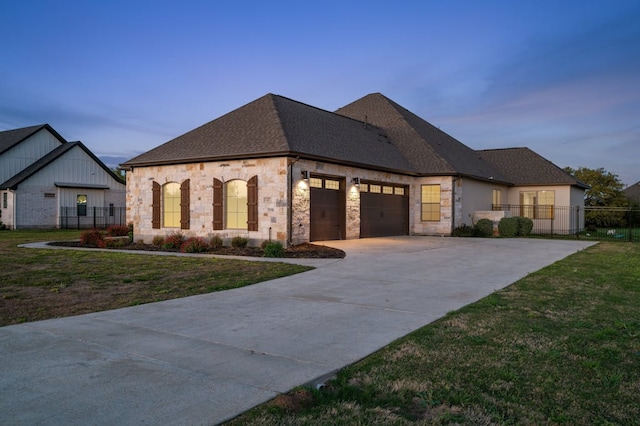 french country inspired facade with a garage, a shingled roof, fence, driveway, and a lawn