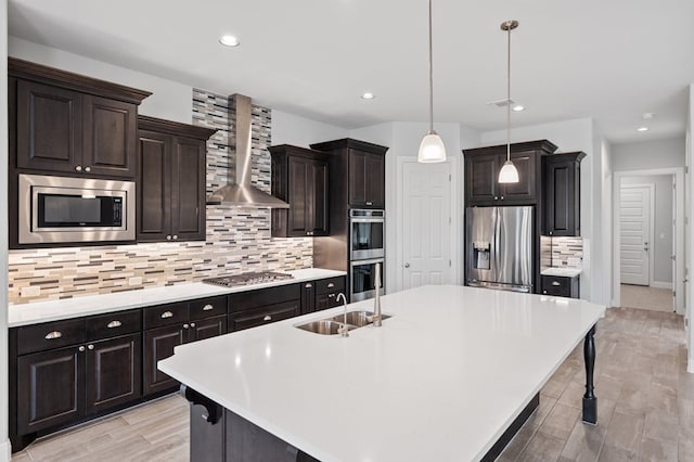 kitchen featuring a sink, light countertops, appliances with stainless steel finishes, wall chimney range hood, and a center island with sink