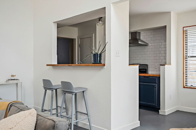 kitchen featuring a breakfast bar area, blue cabinetry, tasteful backsplash, wall chimney range hood, and baseboards