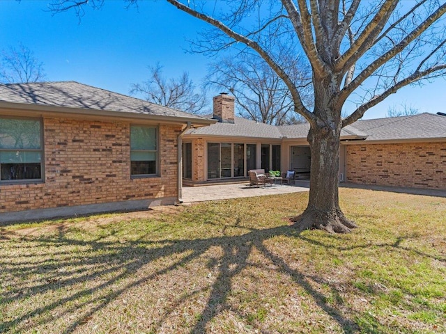 back of house featuring a lawn, roof with shingles, brick siding, a chimney, and a patio area