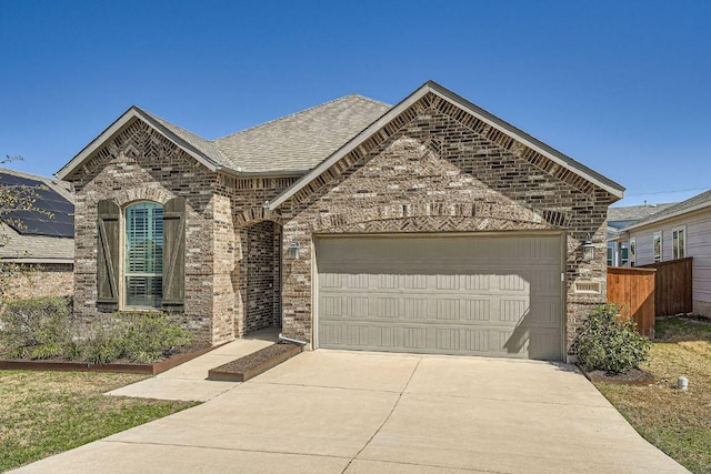 french country home with concrete driveway, roof with shingles, an attached garage, fence, and brick siding