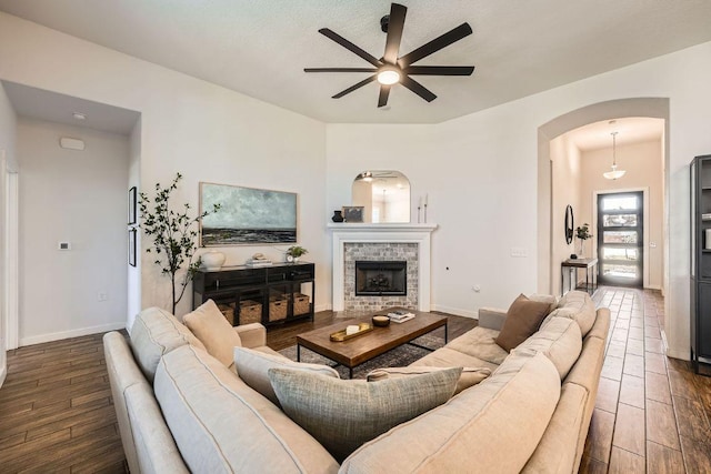 living area featuring baseboards, arched walkways, ceiling fan, dark wood-type flooring, and a brick fireplace