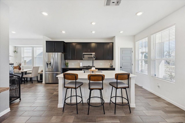 kitchen with visible vents, a kitchen island with sink, backsplash, appliances with stainless steel finishes, and light countertops