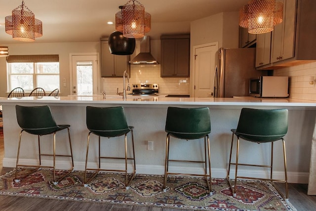 kitchen featuring wall chimney exhaust hood, light countertops, appliances with stainless steel finishes, wood finished floors, and a kitchen bar