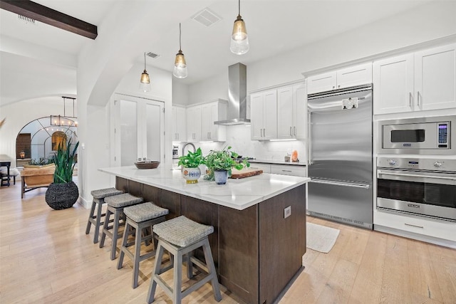 kitchen featuring light wood finished floors, visible vents, decorative backsplash, wall chimney range hood, and built in appliances