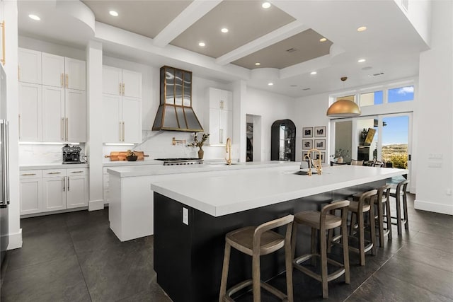 kitchen with premium range hood, white cabinetry, light countertops, backsplash, and a large island with sink