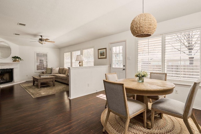 dining area with a fireplace with raised hearth, ceiling fan, visible vents, baseboards, and dark wood finished floors