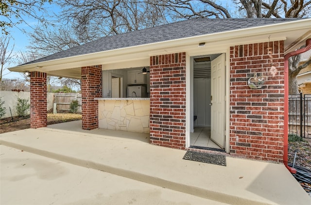 property entrance with a shingled roof, fence, and brick siding