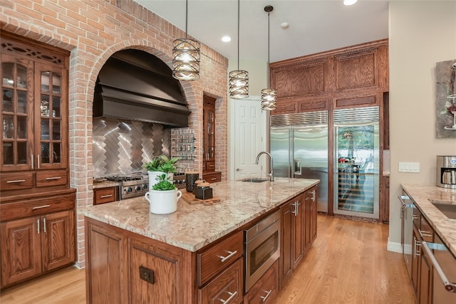 kitchen with brown cabinets, a sink, built in appliances, light wood-type flooring, and premium range hood